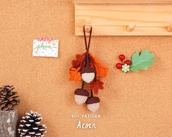 May include: A handmade crochet acorn ornament with brown, orange, and cream colors, hanging from a wooden peg. The ornament is accompanied by a decorative leaf and berries. The text "PDF PATTERN Acorn" is visible.