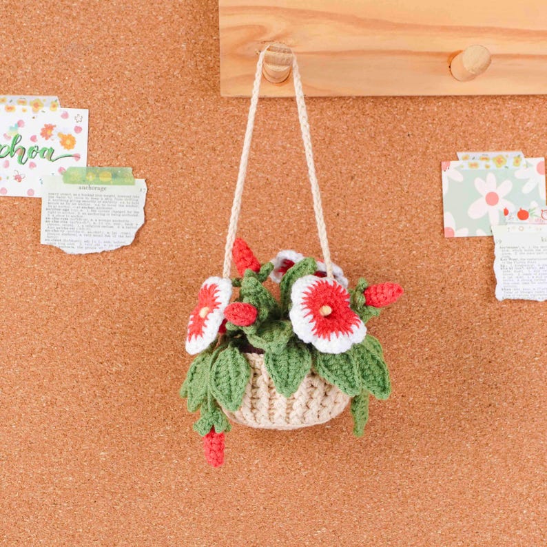 May include: A handmade crochet hanging basket with red and white flowers and green leaves. The basket is beige and suspended by a white cord. The background is a corkboard with decorative paper notes.