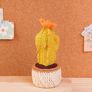May include: A crocheted cactus in a small pot. The cactus is yellow-green with small orange accents and a bright orange flower on top. It sits in a dark brown soil base inside a textured, off-white crocheted pot with a light brown rim at the top.