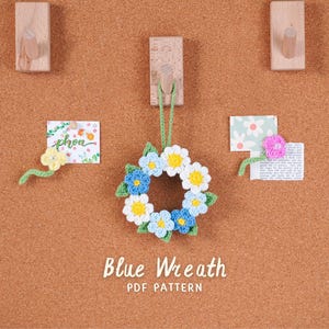 Peut inclure: Une couronne au crochet faite à la main avec des détails de fleurs bleues, blanches et jaunes, suspendue sur un fond en liège. Le texte "Blue Wreath PDF Pattern" est affiché en dessous. Des accents floraux décoratifs sont également épinglés.