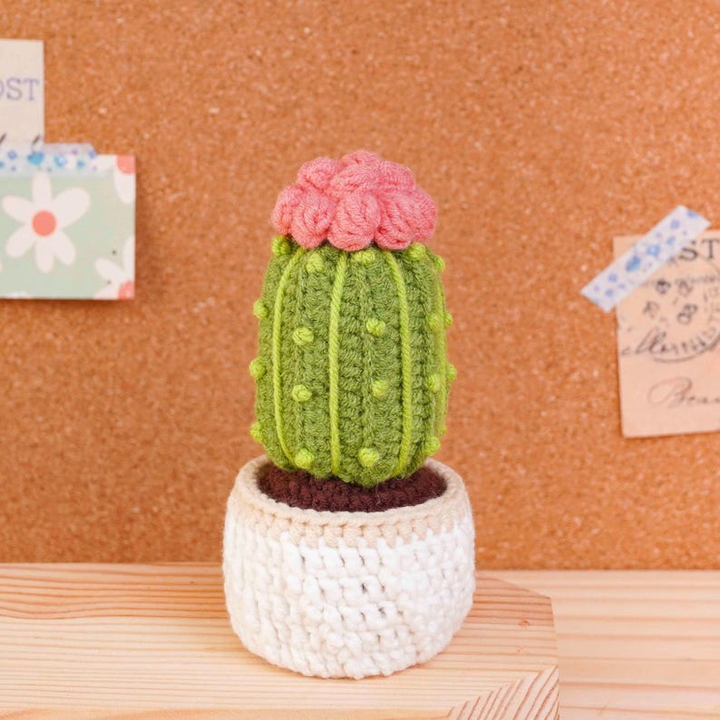 May include: A crocheted cactus with a bright green body featuring vertical ridges and small yellow dots, topped with a cluster of pink flowers. The cactus is planted in a white crocheted pot with a beige rim, showcasing detailed yarn texture.