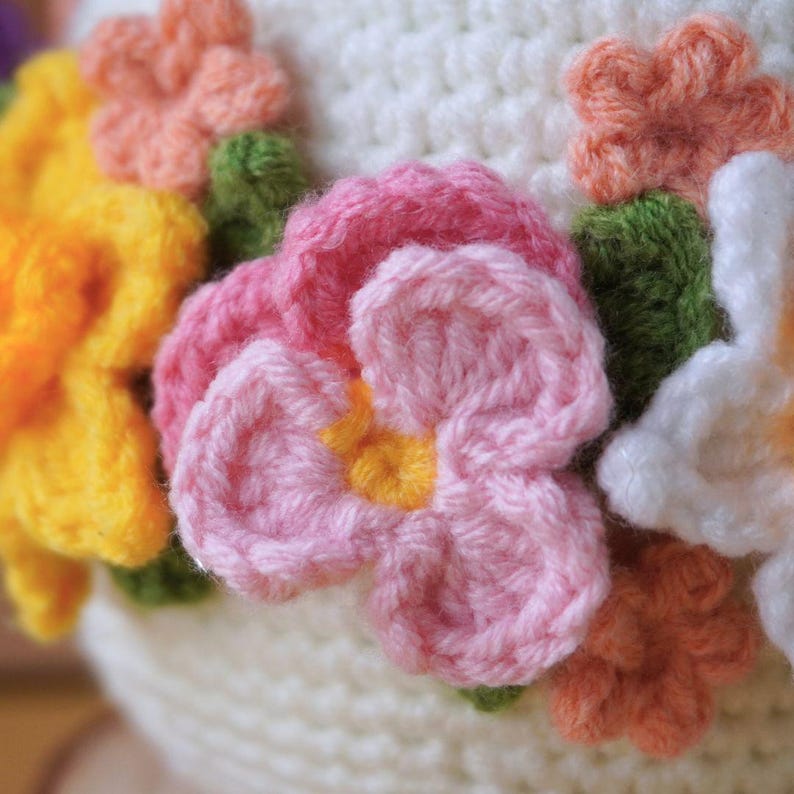May include: A white crocheted hat with a variety of crocheted flowers in pink, yellow, orange, and white.