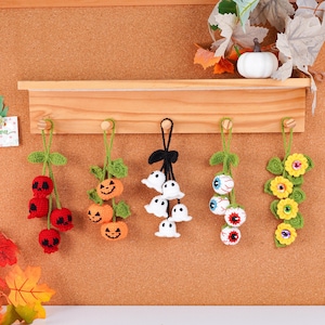 May include: A collection of handmade Halloween ornaments. The ornaments include red skull cherries, orange jack-o'-lanterns, white ghost clusters, eyeball clusters, and yellow flower clusters. Each ornament is crocheted and hangs from a green stem.