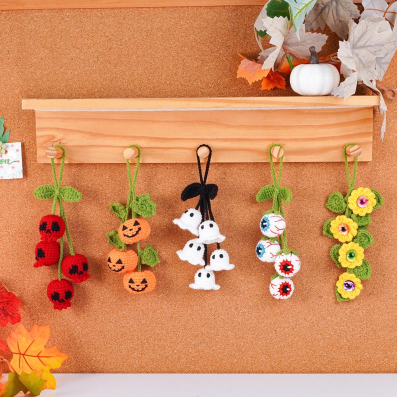 May include: A collection of handmade Halloween ornaments. The ornaments include red skull cherries, orange jack-o'-lanterns, white ghost clusters, eyeball clusters, and yellow flower clusters. Each ornament is crocheted and hangs from a green stem.