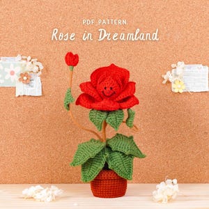 Pode incluir: Uma rosa vermelha de crochê com um rosto sorridente, folhas verdes e um caule marrom, plantada em um pequeno vaso cor de terracota. O texto "Rose in Dreamland" está acima da rosa. O fundo é uma placa de cortiça.