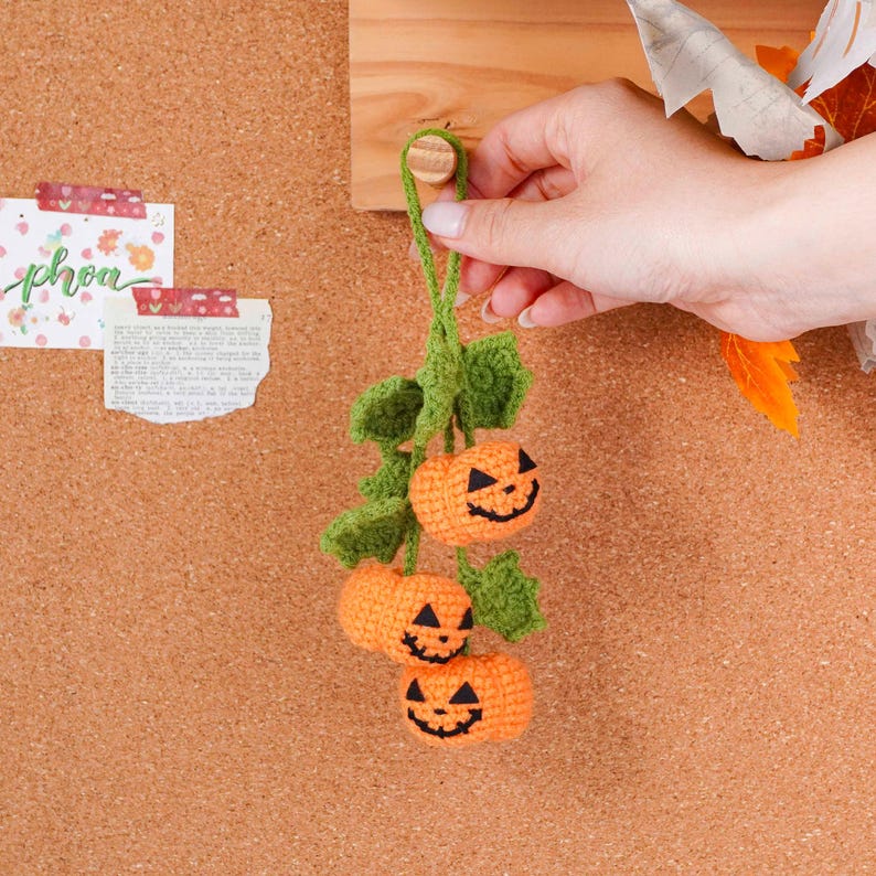 May include: A handmade crochet Halloween decoration featuring three orange pumpkins with black embroidered jack-o'-lantern faces, connected by green leaves and a green stem. The pumpkins are hanging from a wooden peg.