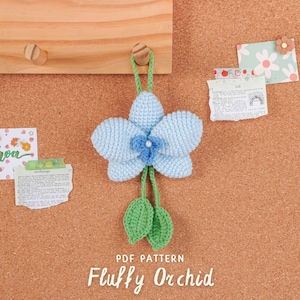 Peut inclure: Une orchidée crochetée bleu clair, avec un centre bleu plus foncé et des feuilles vertes, suspendue à une cheville en bois. Le texte "PDF PATTERN" et "Fluffy Orchid" sont en bas. Des notes décoratives sont épinglées sur le fond en liège.