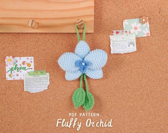 May include: A crocheted orchid in light blue, with a darker blue centre and green leaves, suspended from a wooden peg. The text "PDF PATTERN" and "Fluffy Orchid" are at the bottom. Decorative notes are pinned to the corkboard background.