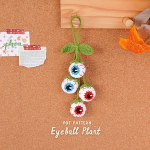 Könnte beinhalten: Eine handgefertigte gehäkelte Augapfelpflanze mit vier weißen, roten und blauen Augäpfeln, die an einem grünen Stiel und Blättern hängen. Der Text "PDF PATTERN" und "Eyeball Plant" befindet sich unten. Der Hintergrund ist eine Pinnwand mit dekorativen Notizen.