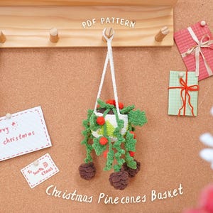 Könnte beinhalten: Ein handgefertigter Weihnachts-Kiefernzapfen-Korb, gehäkelt mit grünen Blättern, roten Beeren und braunen Kiefernzapfen, hängt an einem weißen Kordel. Das Bild zeigt eine Holztafel mit dem Text "PDF PATTERN" und Weihnachtskarten.