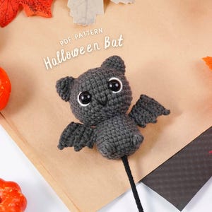 Può includere: Un pipistrello di Halloween grigio all'uncinetto con grandi occhi bianchi e piccole ali. Il pipistrello è su un bastoncino nero ed è circondato da foglie d'autunno e zucche. Il testo "PDF PATTERN Halloween Bat" è visibile.