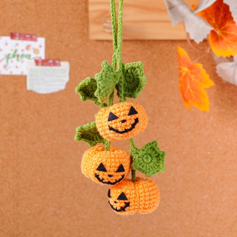 May include: A handmade Halloween decoration featuring three crocheted orange pumpkins with black smiling faces and green leaves. The pumpkins are strung together with green yarn, creating a festive autumn ornament.