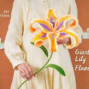 Op de afbeelding: Een grote, gehaakte leliebloem in tinten geel, oranje en paars, vastgehouden door een persoon die een crèmekleurige jurk draagt. De bloem heeft groene bladeren en een stengel. Tekst op de afbeelding luidt "PDF PATTERN" en "Giant Lily Flower".