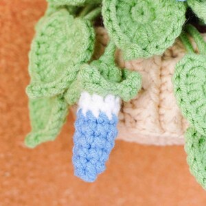 May include: Close-up of a crocheted plant with green leaves and a light beige pot. The plant features a blue and white crocheted flower. The leaves are a vibrant green, and the pot has a textured appearance.