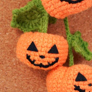 May include: Close-up of crocheted orange pumpkins with black triangle eyes and a curved mouth. Green leaves and stems are attached to the pumpkins. The pumpkins are likely for Halloween decorations.