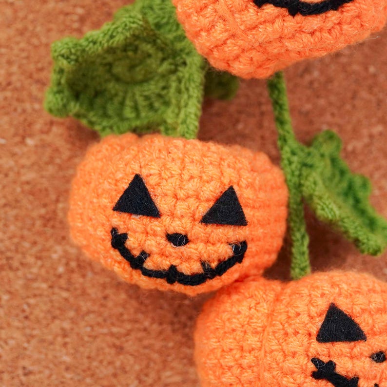 May include: Close-up of crocheted orange pumpkins with black triangle eyes and a curved mouth. Green leaves and stems are attached to the pumpkins. The pumpkins are likely for Halloween decorations.