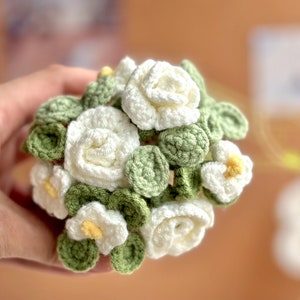 Op de afbeelding: Een gehaakt boeket van witte bloemen met groene bladeren. De bloemen zijn in een cirkelvorm gerangschikt en worden in een hand gehouden.