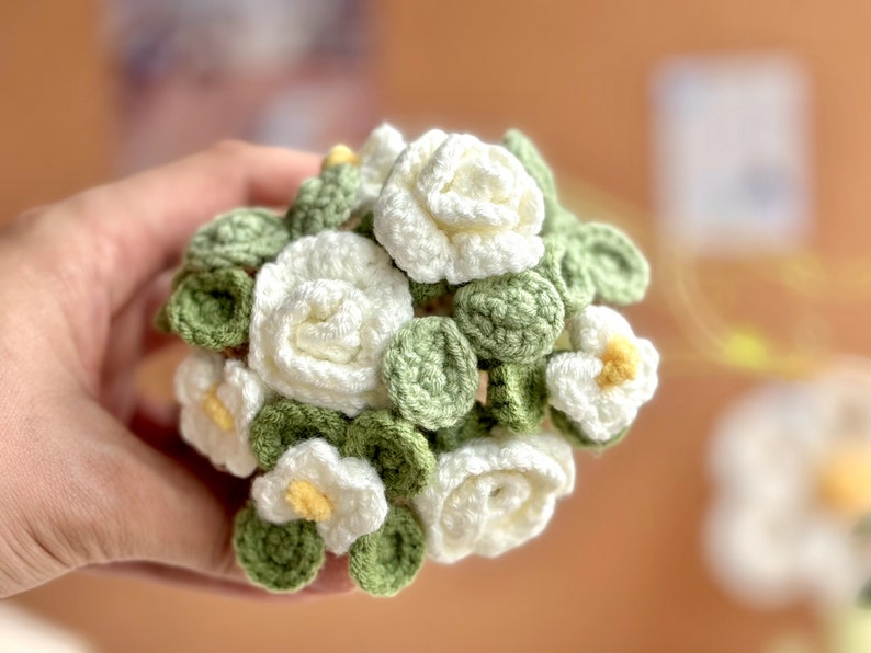 Op de afbeelding: Een gehaakt boeket van witte bloemen met groene bladeren. De bloemen zijn in een cirkelvorm gerangschikt en worden in een hand gehouden.