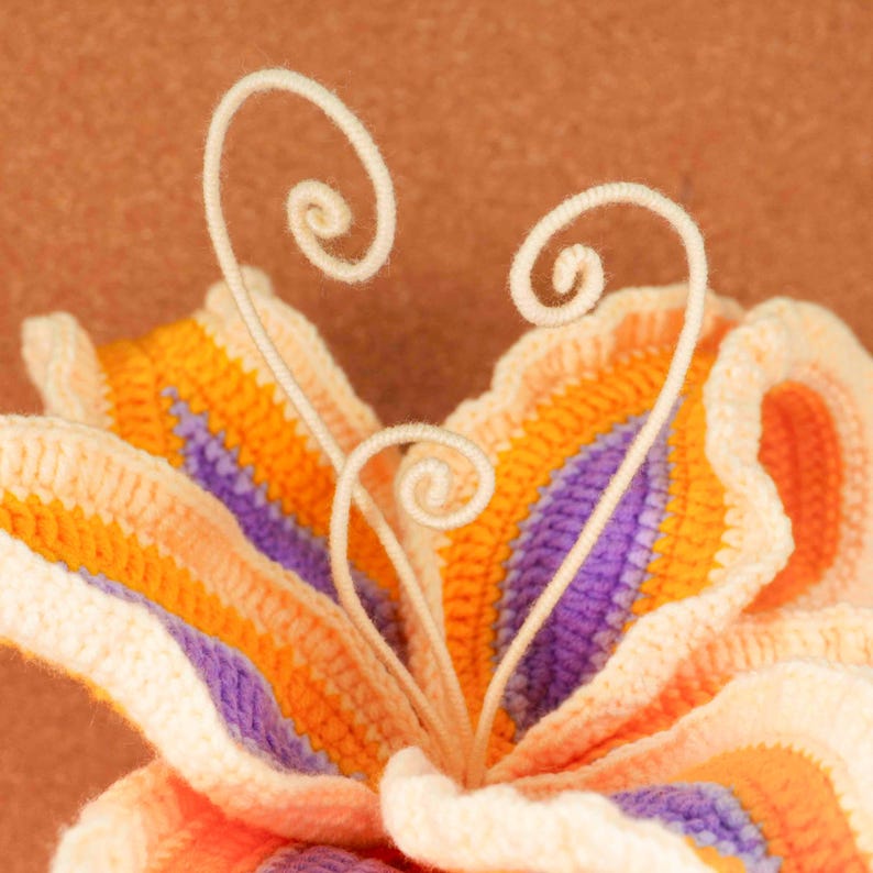 Op de afbeelding: Close-up van een handgemaakte gehaakte bloem in tinten oranje, paars en cr&egrave;me. De bloem heeft een gedetailleerd, getextureerd ontwerp met gekrulde, cr&egrave;mekleurige accenten. De bloemblaadjes zijn gelaagd, waardoor een driedimensionaal effect ontstaat.