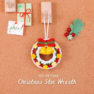 以下が含まれることがあります： 赤と緑のリボン、黄色のベル、赤と黄色の星のアクセントが付いた、手作りのクリスマスの星のリース。白い紐から吊るされています。「Christmas Star Wreath」の文字が下にあります。