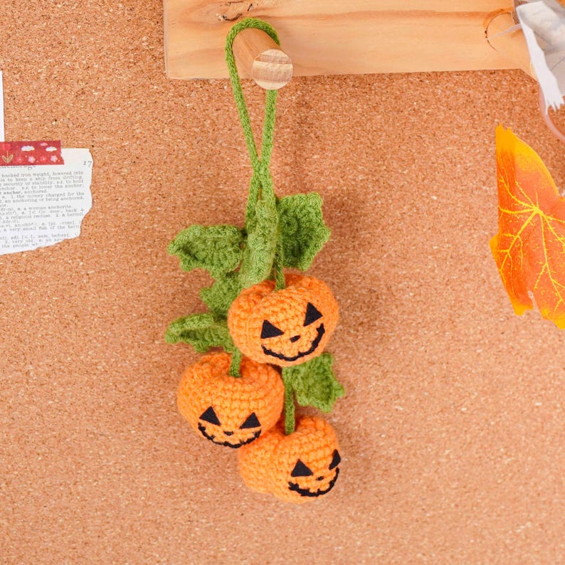 May include: A handmade crochet Halloween decoration featuring three orange pumpkins with black jack-o'-lantern faces. The pumpkins are attached to green leaves and a green stem, perfect for seasonal decor. The decoration is hanging from a wooden peg.