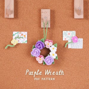 May include: A crocheted floral wreath in shades of purple, pink, and white, suspended against a corkboard. The image includes the text "Purple Wreath PDF Pattern". The wreath is a decorative item.