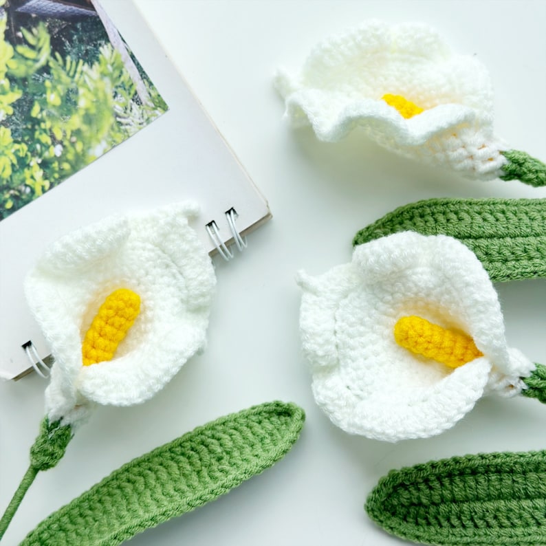 May include: Three white crocheted calla lily flowers with yellow centers and green crocheted leaves.
