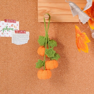May include: A handmade crochet pumpkin garland with three orange pumpkins and green leaves. The garland is hanging against a corkboard background, evoking a fall or autumn theme. The pumpkins are approximately 2 inches in diameter.