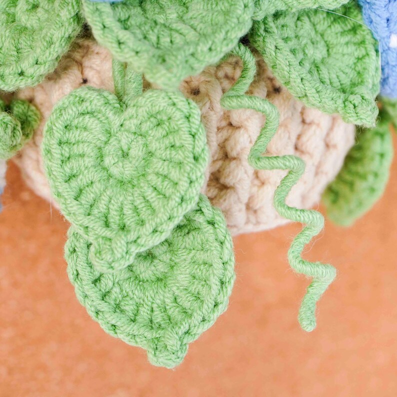 May include: Close-up of a handmade crochet plant with green leaves and a beige pot. The leaves are heart-shaped and the pot has a textured appearance. A green, curly vine adds detail to the design. The craftwork is set against a soft orange background.