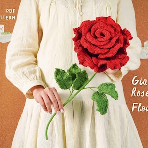 Puede incluir: Una gran rosa roja hecha a mano con ganchillo, con hojas y tallo verdes. Las palabras "Giant Rose Flower" son visibles en la imagen. La rosa se sostiene contra un vestido color crema.