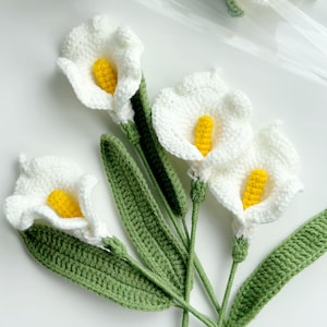May include: A bouquet of crocheted white calla lilies with yellow centers. The lilies are arranged with green crocheted leaves.