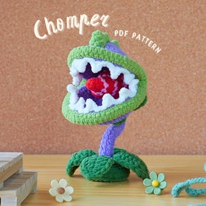 Puede incluir: Un juguete de peluche tejido a crochet de una planta Chomper del videojuego Plants vs. Zombies. El juguete es verde, morado y blanco y tiene una boca grande abierta con una lengua roja. El juguete está sentado en una mesa de madera con un fondo marrón. El texto "Chomper PDF PATTERN" está escrito encima del juguete.