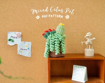 May include: A crocheted mixed cactus pot with a brown pot and green and yellow cacti. The pot is sitting on a wooden shelf with a calendar and a wooden figurine.