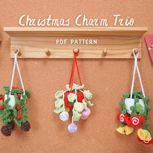 Puede incluir: Tres adornos de plantas colgantes de ganchillo con temas navideños. Los adornos cuelgan de ganchos en una estantería de madera. La estantería tiene el texto "Christmas Charm Trio" y "PDF Pattern" escrito en ella.