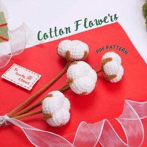 Könnte beinhalten: Ein Set aus drei weißen Baumwollblumen-Häkelmustern auf rotem Hintergrund mit weißem Band und einem Zettel mit der Aufschrift "To Santa, He Claus". Der Text "Cotton Flowers PDF Pattern" ist in grünen Buchstaben auf dem roten Hintergrund geschrieben.