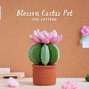 May include: Crocheted cactus pot with a pink flower on top. The pot is brown and the cactus is green. The text "Blossom Cactus Pot PDF Pattern" is written above the cactus.
