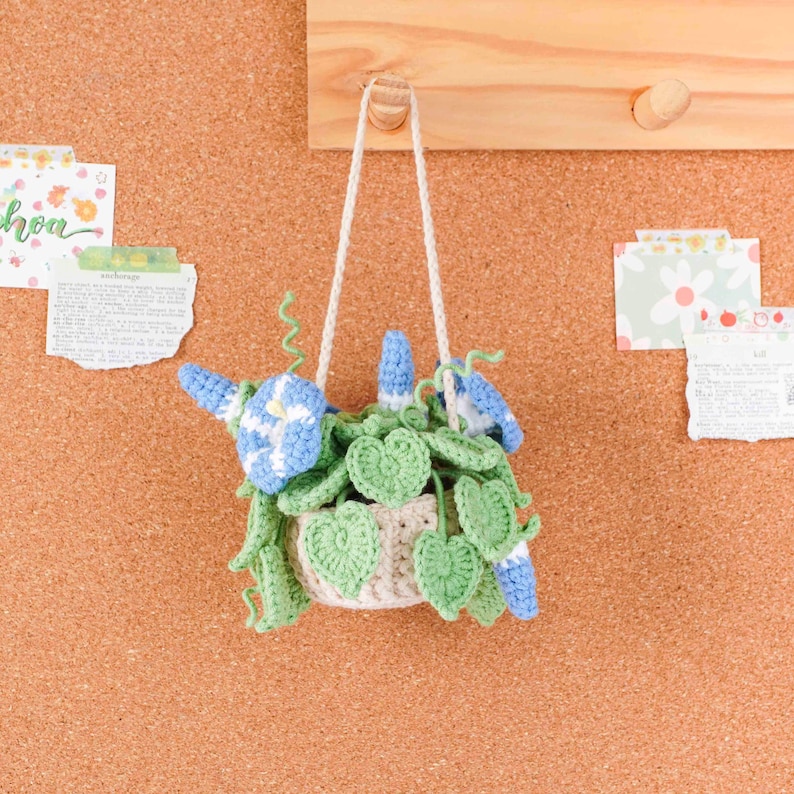 May include: A handmade crochet hanging planter with a cream-colored pot, green leaves, and blue and white flowers. The planter is suspended by a white cord and is displayed against a corkboard background.