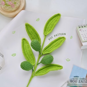 Op de afbeelding: Een gehaakte olijftak met groene bladeren en olijven. De tak ligt op een wit oppervlak met de tekst "PDF PATTERN" erop.