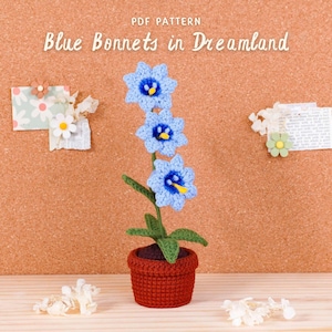 Op de afbeelding: Een gehaakte blauwe bloemenplant in een rode pot. De plant heeft drie blauwe bloemen met gele centra en groene bladeren. De tekst "Blue Bonnets in Dreamland" staat bovenaan. De achtergrond is een prikbord.