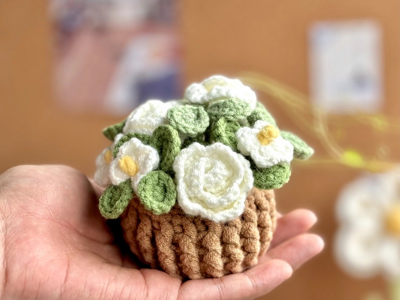 Op de afbeelding: Een gehaakte bloempot met witte bloemen en groene bladeren. De pot is bruin en heeft een textuur.
