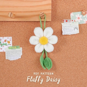 May include: A crocheted daisy in white and yellow, with green leaves, suspended on a corkboard. The text "Fluffy Daisy" is below the flower. Several small notes and decorative items are also pinned to the board, creating a charming display.