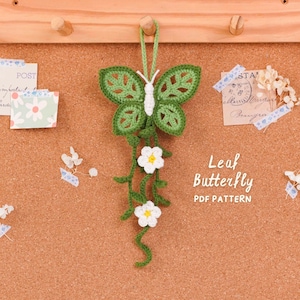 May include: A crocheted green butterfly ornament with a white body and two white flowers hanging from green vines. The text Leaf Butterfly PDF PATTERN is printed in white on the right side of the object against a cork board background.