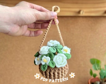 May include: A crocheted hanging planter with white flowers and green leaves. The planter is brown and has a rope handle. The text "PDF PATTERN" is at the bottom of the image.