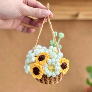 Op de afbeelding: Een gehaakte hangende plantenhanger met een bruine mand gevuld met witte en gele bloemen. De hanger hangt aan een bruin touw.