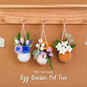 Peut inclure: Trois pots de fleurs suspendus en forme d'œuf au crochet avec des fleurs colorées. Les pots sont suspendus à une étagère en bois. Le texte "Egg Garden Pot Trio" est affiché en dessous. Le fond est un tableau en liège.
