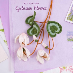 May include: A crocheted cyclamen flower pattern with the text "PDF PATTERN Cyclamen Flower" in a handwritten style. The flower is white and pink, with green and white leaves. The background is a light purple.