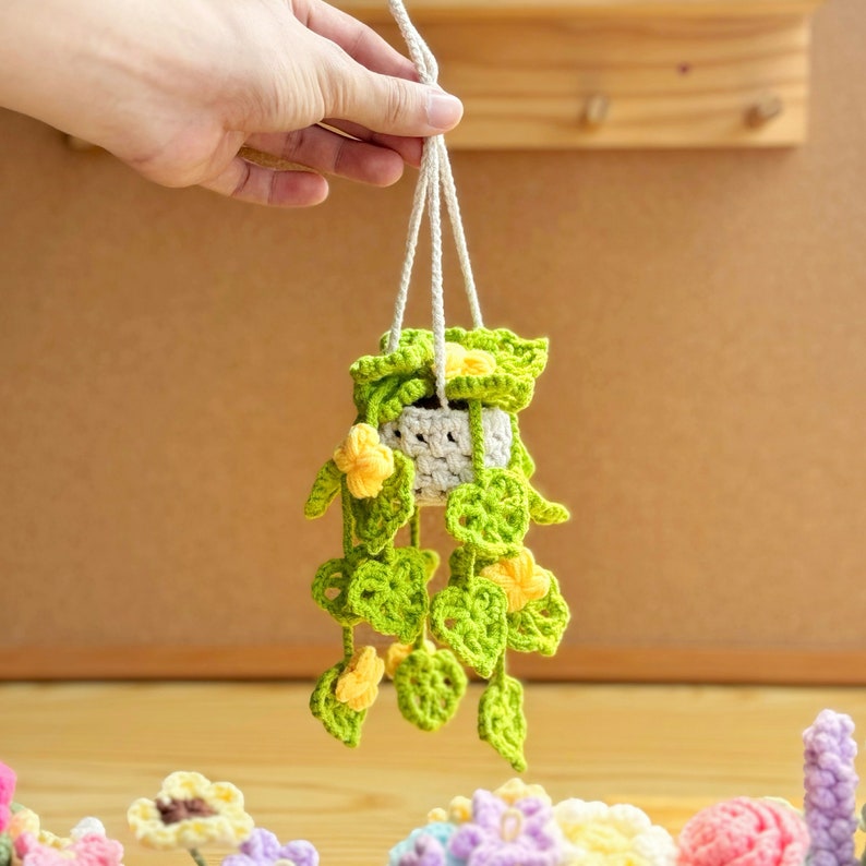 May include: A crocheted hanging planter with green leaves and yellow flowers. The planter is white and has a crocheted design. The planter is hanging from a white string.