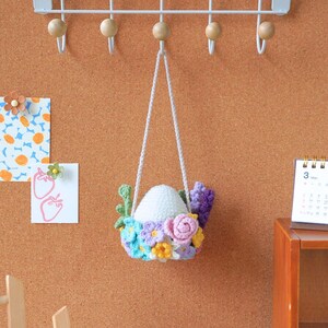 May include: A white crocheted egg with a floral wreath hanging from a white string. The wreath is made of pink, purple, blue, and yellow flowers.