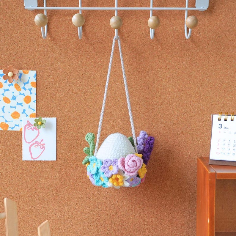 May include: A white crocheted egg with a floral wreath hanging from a white string. The wreath is made of pink, purple, blue, and yellow flowers.