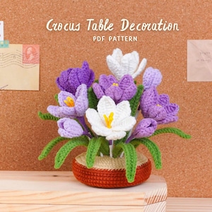May include: A crocheted floral arrangement featuring purple and white crocus flowers with yellow centers, green stems, and leaves. The flowers are in a small, round, orange pot. The text "Crocus Table Decoration PDF Pattern" is above the flowers.